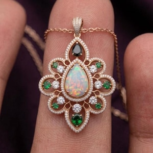 May include: A rose gold pendant featuring a central pear-shaped opal surrounded by a floral-inspired frame of small round white diamonds and green gemstones. A single black teardrop stone sits at the top, all set on a delicate rose gold chain.