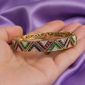 Op de afbeelding: Een scharnierende gouden armband met een herhalend geometrisch chevronpatroon. Het ontwerp is ingelegd met vierkant geslepen smaragden, robijnen, saffieren en kleine ronde diamanten die in afwisselende driehoekige secties over de band zijn geplaatst.
