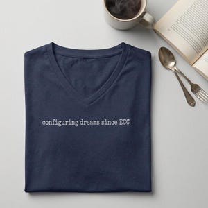 May include: A navy blue V-neck t-shirt featuring the white text "configuring dreams since ECC" printed across the chest. The shirt is folded neatly, showcasing the neckline and the short sleeves, with the text centered in a typewriter-style font.