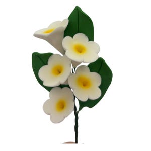 May include: A decorative floral spray featuring five white flowers with bright yellow centers and several green leaves. The flowers are arranged on a thin, dark green stem, creating a cluster of blossoms that are attached to a single wire base.