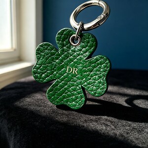 May include: A four-leaf clover shaped keychain made of textured green leather with dark green stitching along the edges. The center features the gold embossed letters DR. A silver-toned metal split ring is attached to the top via a metal eyelet.