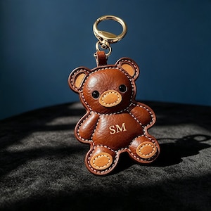 Può includere: Un portachiavi in pelle marrone a forma di orsetto con un anello in metallo dorato, dettagli color cuoio su orecchie, muso e zampe, cuciture bianche a contrasto e le lettere SM impresse in oro al centro del busto dell'orsetto.