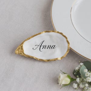 May include: A white oyster shell dish with a gold-painted rim and the name Anna printed in black script in the centre. The shell is positioned next to a white plate with a gold rim and a small cluster of white flowers with green leaves.