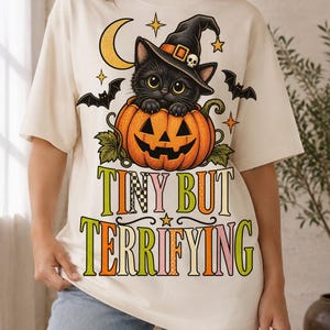May include: An off-white t-shirt featuring a graphic of a black cat wearing a witch hat inside a carved orange pumpkin. The design includes bats, stars, a crescent moon, and the text "TINY BUT TERRIFYING" in colorful, patterned lettering.