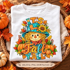 Può includere: Una t-shirt bianca con una grafica colorata di uno spaventapasseri con cappello blu, circondato da girasoli e zucche. Il testo IT'S FALL Y'ALL è scritto in un carattere decorativo. Il design include foglie autunnali colorate.