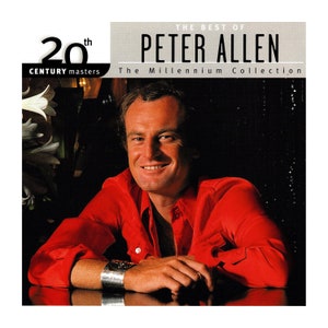 May include: A red shirt with a button-up collar and a silver bracelet. The man in the photo is smiling and has brown hair. The text on the album cover reads "The Best of Peter Allen The Millennium Collection 20th Century Masters".