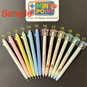 May include: A collection of twelve retractable pens numbered 10 through 21. The pens feature pastel-colored barrels, some with animal-themed toppers or small house-shaped caps. A white sign at the top displays the text Mint Point Gaming.