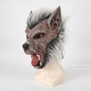 May include: A werewolf mask featuring a brown textured face with a black nose, an open mouth revealing white teeth, and a red tongue. The mask is accented with long, shaggy white and black faux fur around the ears and sides of the head.