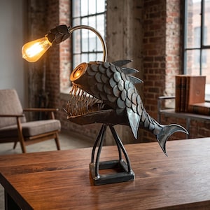 Puede incluir: Una lámpara de mesa de metal con forma de pez rape en acabado gris oscuro, con placas superpuestas en forma de escamas, una mandíbula con dientes afilados y un brazo flexible que sostiene una bombilla de estilo Edison brillante.