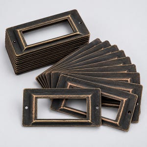 May include: A collection of rectangular metal label holders featuring a dark bronze finish with distressed highlights. Each frame has a central rectangular cutout and two small circular screw holes on the left and right sides for mounting purposes.