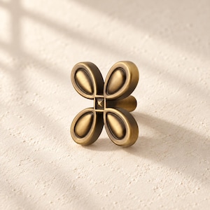 May include: A brass-toned metal cabinet knob featuring a four-petaled flower design. The center of the knob has a small square detail with a raised point, and each petal has a smooth, rounded, recessed surface with a darker metallic finish.