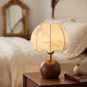 May include: A table lamp featuring a spherical dark wood base and a brass stem. The lamp has a scalloped, off-white fabric shade decorated with delicate, thin-lined botanical illustrations of leaves and branches in a dark ink color.
