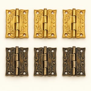 May include: Six rectangular metal hinges arranged in two rows of three. The top row features three bright gold-toned hinges, while the bottom row contains three dark bronze-toned hinges. Each hinge has a central barrel and four screw holes.