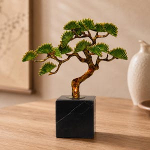 May include: A decorative sculpture of a tree with a metallic gold-toned trunk and branches, featuring fan-shaped green leaves. The tree is mounted on a solid, square black marble base with visible white veining throughout the stone surface.