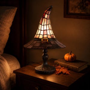 May include: A stained glass table lamp shaped like a pointed witch hat with a dark brim and a lighter, multi-toned crown. The lamp features a metallic base with a fluted design and emits a warm glow through the glass panels.