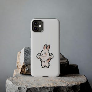 May include: A white smartphone case featuring a central graphic illustration of a small, muscular rabbit with defined arms and chest. The rabbit has pink-tinted ears and cheeks, and a small cross symbol on its torso. The case has a dual-lens camera cutout.