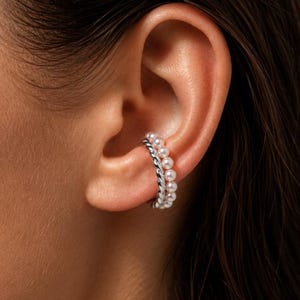 May include: A silver-toned ear cuff featuring a dual-band design. One band consists of a smooth, twisted metal rope texture, while the adjacent band is lined with a row of small, round, white pearls that follow the curve of the ear cartilage.