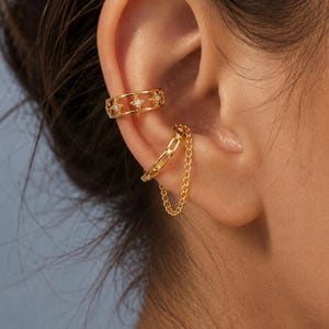 May include: Two gold-toned ear cuffs worn on the cartilage. The upper cuff features a cut-out star pattern with small embedded stones. The lower cuff is a smooth band connected to a delicate dangling chain that loops back to the ear.