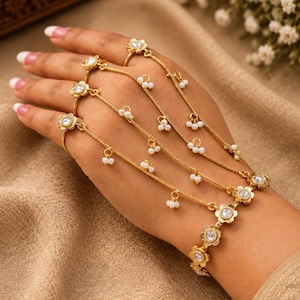 May include: A gold-toned hand chain bracelet featuring a series of connected flower-shaped links with central clear stones. The design includes multiple delicate chains extending from a wrist band to individual rings, adorned with small white pearls.