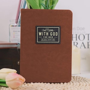Puede incluir: Un libro rectangular de color marrón con textura de cuero y una etiqueta rectangular negra en la portada. La etiqueta presenta el texto blanco "One Minute WITH GOD FOR MEN 365 DAILY DEVOTIONS" con líneas decorativas encima y debajo del título.