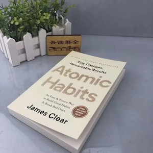 May include: A paperback book titled Atomic Habits by James Clear with a cream cover and gold lettering. The subtitle reads Tiny Changes, Remarkable Results. An Easy & Proven Way to Build Good Habits & Break Bad Ones. A brown circle states Over 1 Million Copies Sold.
