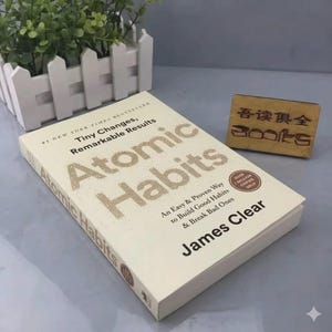 May include: A copy of the book Atomic Habits by James Clear with a cream-colored cover featuring gold-toned text. The book title is prominently displayed alongside the subtitle Tiny Changes, Remarkable Results and An Easy & Proven Way to Build Good Habits & Break Bad Ones. A small wooden sign with Chinese characters and the word BOOKS is nearby.
