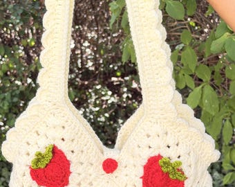 May include: A cream-colored crochet tote bag featuring a repeating granny square pattern. The bag is decorated with several red strawberry appliqués, each topped with small green leaves. It includes a single wide, scalloped-edge shoulder strap.