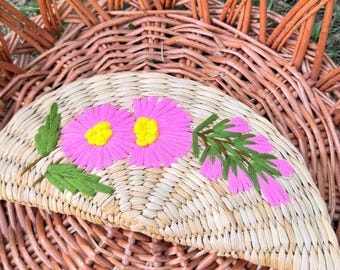 May include: A semi-circular woven straw clutch featuring embroidered pink flowers with yellow centers and green leaves, resting inside a large, round, brown wicker basket with a woven base and arched, radiating side supports.