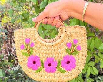 May include: A semi-circular woven straw handbag featuring two round wooden handles. The front of the bag is decorated with embroidered bright pink flowers and green leaves arranged in a horizontal pattern across the center of the natural tan straw.