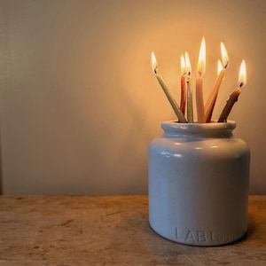 May include: A cylindrical white ceramic jar with the embossed text LAB Lagoon on the base, containing several thin, lit taper candles in various muted earth tones. The candles have visible flames, casting a warm glow from within the container.