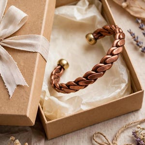 May include: A copper-toned metal cuff bracelet featuring a braided design with smooth, rounded gold-toned end caps. The bracelet is presented inside an open, square-shaped cardboard gift box lined with light-coloured tissue paper.