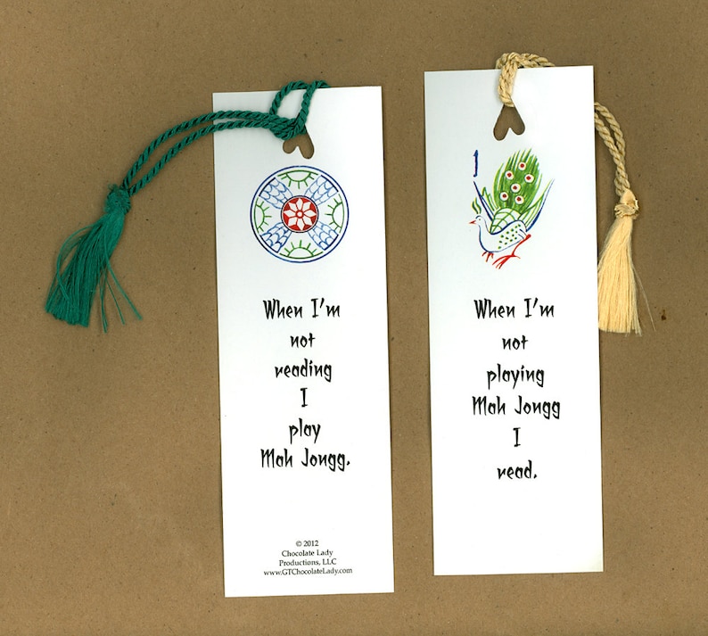 May include: Two white bookmarks with green and white tassels. The bookmark on the left has a blue and green circular design with a red flower in the center. The bookmark on the right has a green and white peacock design. Both bookmarks have text that reads "When I'm not reading I play Mah Jongg." and "When I'm not playing Mah Jongg I read."
