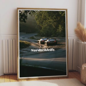 Puede incluir: Un cuadro vertical enmarcado que muestra dos coches de carreras en una pista de asfalto sinuosa a través de un bosque. La imagen presenta el texto Nordschleife en blanco, centrado sobre la carretera. El marco es de madera clara.