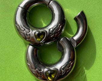 May include: Two silver-toned circular body jewelry pieces featuring a hinged segment design. Each piece is engraved with flame patterns and set with a single heart-shaped olive green gemstone at the center of the curved metal surface.