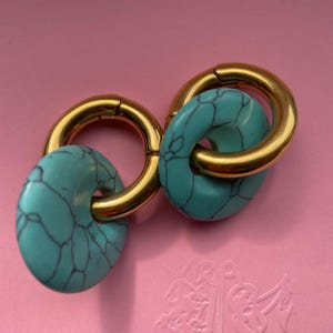 May include: A pair of hoop earrings featuring polished gold-tone metal rings. Each ring is threaded through a circular, turquoise-colored stone bead with a dark, web-like veining pattern throughout the surface of the stone material.