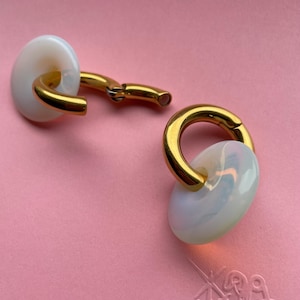 May include: A pair of hoop earrings featuring a polished gold-tone metal band and a smooth, translucent white circular charm. The earrings have a hinged closure mechanism, allowing the gold hoop to open and close securely through the center hole.