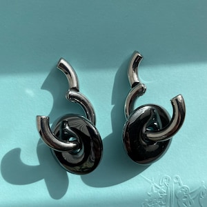 May include: A pair of earrings featuring a polished silver-tone curved metal component that loops through a smooth, circular, dark charcoal-colored disc. The design showcases a high-shine metallic finish with a reflective surface and a modern, sculptural shape.