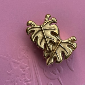 May include: A gold-toned metal brooch shaped like two overlapping monstera leaves. The piece features a textured, matte finish on the leaf surfaces with smooth, raised central veins and defined edges, creating a layered botanical appearance.