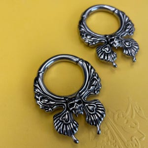 May include: Two silver-toned circular hoop earrings featuring a detailed skull motif at the base, integrated into a wing-like structure with embossed heart shapes and radiating lines. The metal has a polished finish with dark recessed detailing.