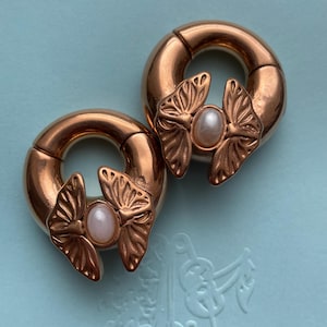 May include: A pair of rose gold-toned circular ear weights featuring a hinged design. Each piece is adorned with a metallic butterfly motif centered with a smooth, oval-shaped, pale pink cabochon stone. The jewelry has a polished finish.