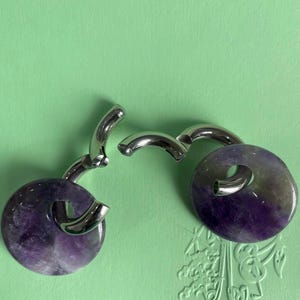 May include: A pair of hoop earrings featuring polished silver-tone metal curved bars that hook through circular, flat, polished amethyst gemstone pendants. The purple stones exhibit natural color variations ranging from deep violet to translucent white.