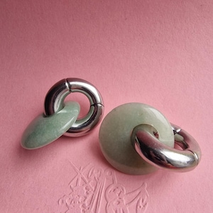 May include: A pair of earrings featuring polished silver-tone metal hoops linked to smooth, circular pale green stone discs. The earrings have a reflective metallic finish and a soft, matte appearance on the stone components, creating a contrast.