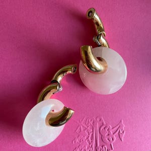 May include: A pair of hoop earrings featuring a polished gold-tone metal curved bar that loops through a circular, translucent pale pink stone disc. The design showcases a smooth metallic finish contrasted with the soft, semi-opaque gemstone element.