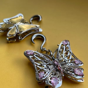 May include: A pair of silver-toned butterfly-shaped ear weights featuring intricate openwork metal detailing. One butterfly is accented with pink teardrop-shaped stones, while the other displays a smooth, polished gold-toned finish on its wings.