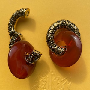 May include: A pair of ear weights featuring a curved, ornate gold-tone metal component with intricate black detailing, connected to a smooth, translucent, deep orange circular ring. One piece is open, while the other is closed in a loop.