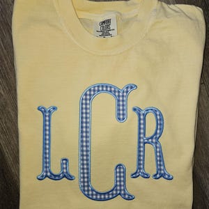 May include: A light yellow Comfort Colors t-shirt with the embroidered letters "LCR" in a blue and white gingham pattern. The shirt is folded on a wooden surface.