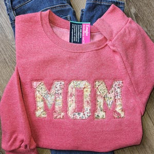 May include: A red sweatshirt with the word "MOM" embroidered across the front in a floral pattern. The sweatshirt is folded on top of a pair of blue jeans. A tag with contact information is attached to the sweatshirt.