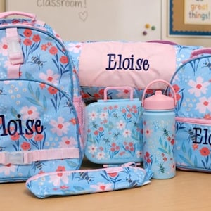May include: A set of school supplies featuring a light blue backpack, a smaller matching bag, a lunch box, a pencil case, and a water bottle, all decorated with a floral and bee pattern. Each item is personalized with the name Eloise in dark blue.