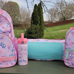 May include: A matching set of school or travel bags in a floral and butterfly pattern. The set includes a backpack, lunch bag, duffel bag, and water bottle. The bags are primarily pink with blue, white, and purple accents.