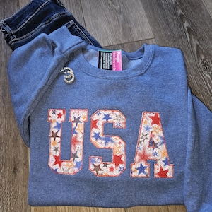 May include: A blue sweatshirt with the word "USA" in a star-patterned design. The sweatshirt is paired with a pair of blue jeans. A small, silver brooch is pinned to the sweatshirt.